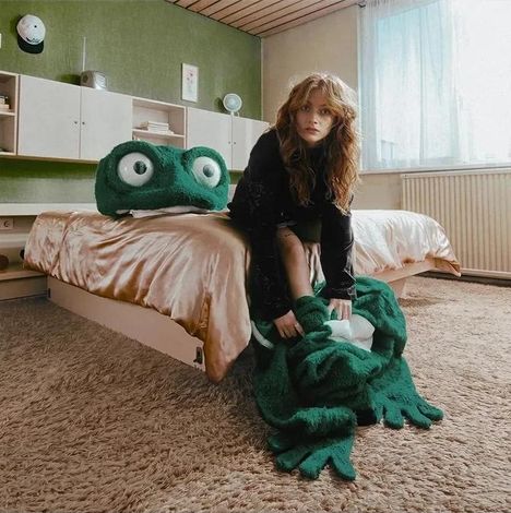 Eine Frau in einem Vintage-Zimmer sitzt auf einem riesigen grünen Plüschfrosch, der auf einem flauschigen Teppich liegt.
