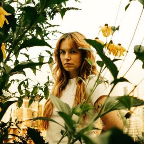 Keine erkennbaren Texte. Eine Frau steht zwischen hohen Pflanzen mit gelben Blüten, schaut direkt, im Hintergrund sind Gebäude.