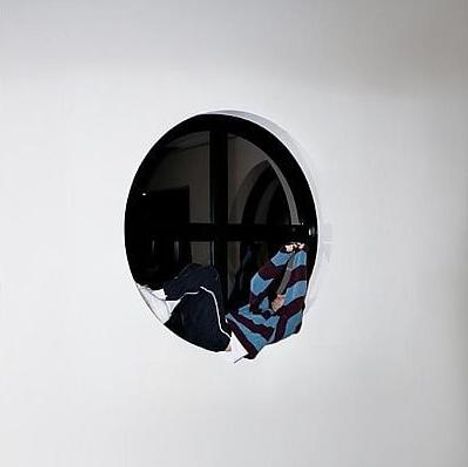 Ein rundes Fenster zeigt einen schwarz-weiß gekleideten Menschen, der sich hineinlehnt.