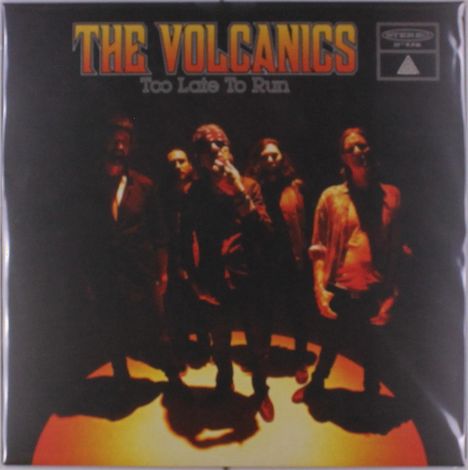 "The Volcanics", "Too Late To Run". Eine Gruppe von Menschen steht im Kreis, in warmem, orangefarbenem Licht.