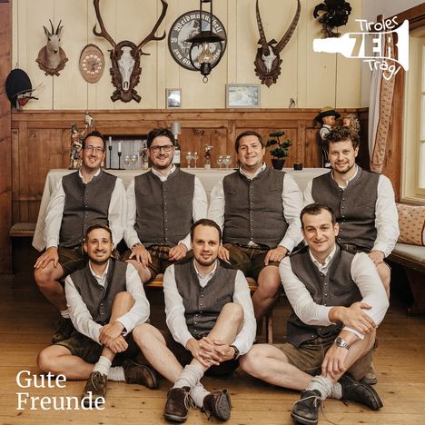 Text: "Gute Freunde, Tiroler ZER Tragl". Sieben Männer in traditioneller Kleidung in einer rustikalen Hütte.