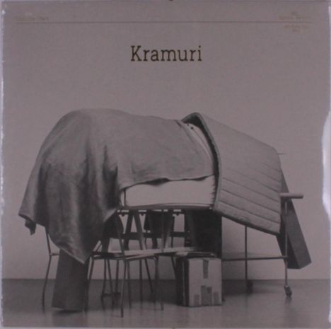 Text: "Kramuri". Eine strukturierte, serpentinenförmige Decke auf mehreren Stapeln von Möbeln und Stühlen.