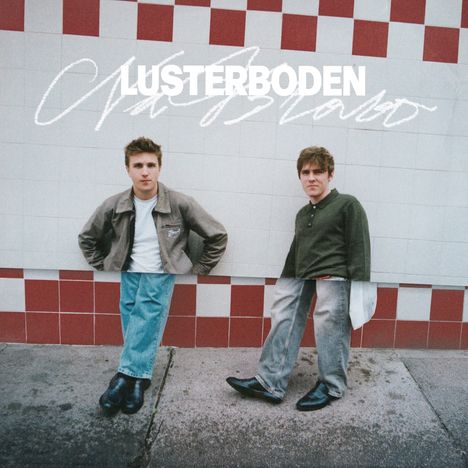 Text: „LUSTERBODEN“. Zwei junge Männer stehen an einer Kachelwand mit rotem Schachbrettmuster am unteren Rand.