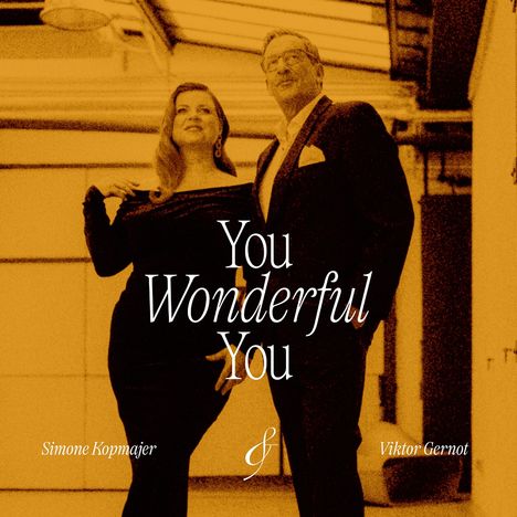 „You Wonderful You“. Simone Kopmajer und Viktor Gernot stehen elegant gekleidet in einem warmen, orangefarbenen Ambiente.