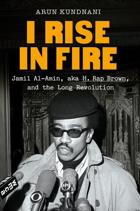 Titel: "I Rise in Fire". Autor: Arun Kundnani. Foto eines Mannes mit Brille und Mütze, umgeben von Mikrofonen.