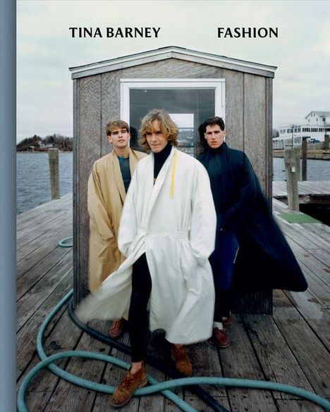 Text: "TINA BARNEY FASHION". Drei Männer in langen Mänteln stehen auf einem Dock vor einem kleinen Holzgebäude, mit Wasser im Hintergrund.