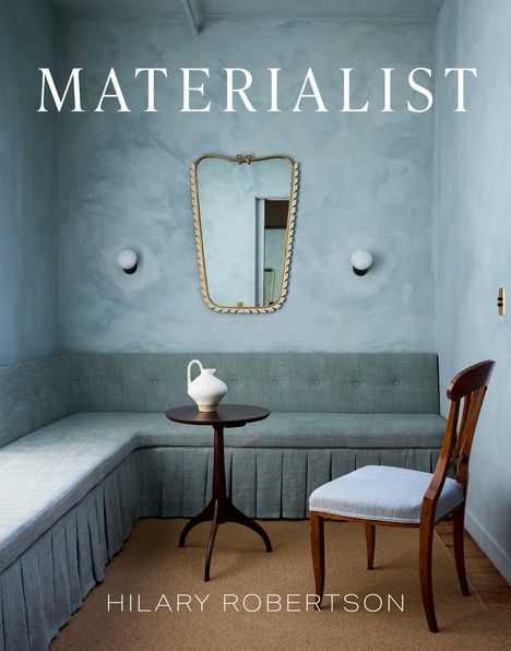 „MATERIALIST“ und „HILARY ROBERTSON.“ Eleganter Raum mit grauem Sofa, Tisch, Stuhl und Spiegel.