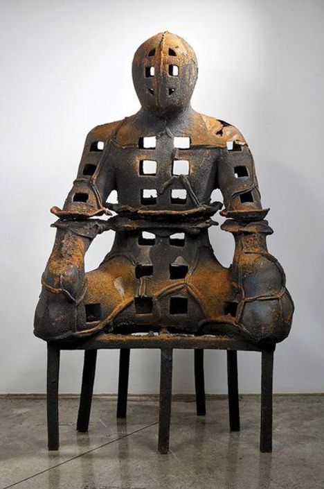 Keine Texte erkennbar. Eine Skulptur mit Löchern, die eine sitzende Figur mit blockartigen Formen darstellt.