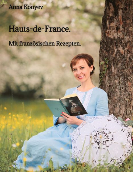 „Anna Konyev: Hauts-de-France. Mit französischen Rezepten.“ Frau im blauen Kleid liest unter Baum mit Sonnenschirm.
