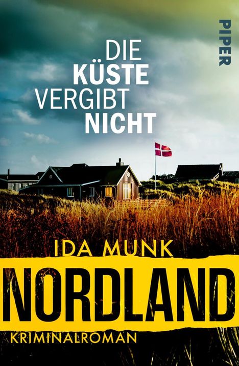 „Die Küste vergibt nicht“, „Ida Munk Nordland Kriminalroman“. Haus und dänische Flagge im Gras, bewölkter Himmel.