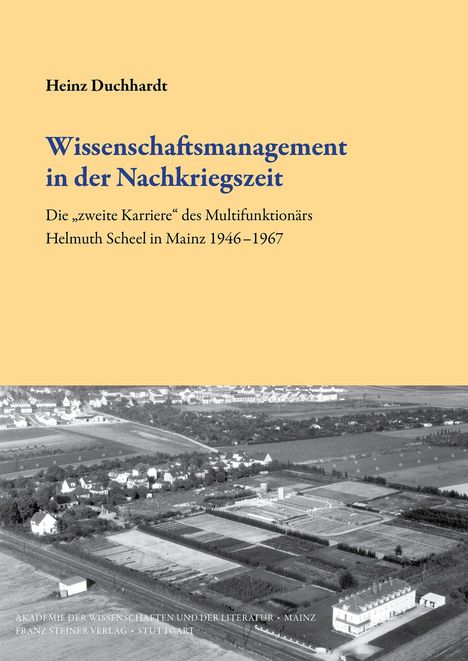 "Wissenschaftsmanagement in der Nachkriegszeit. Luftaufnahme eines ländlichen Gebiets mit Feldern und Gebäuden."