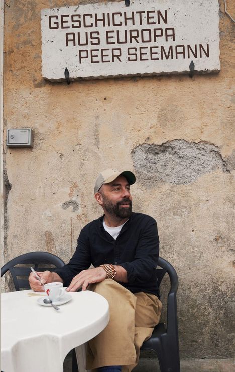 Das Bild zeigt ein Schild mit "GESCHICHTEN AUS EUROPA, PEER SEEMANN". Darunter sitzt ein Mann an einem Tisch mit einer Tasse Kaffee.