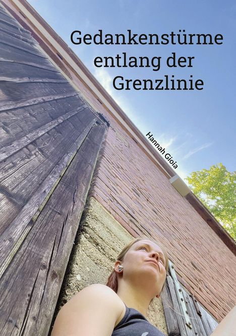 Gedankenstürme entlang der Grenzlinie. Hannah Gioia. Aus ungewöhnlichem Winkel: Person neben alter Holzfassade, blauer Himmel.