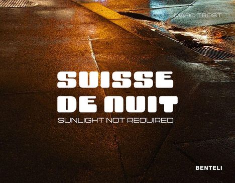 Der Text lautet: "Suisse de Nuit, Sunlight Not Required". Dunkle Straßenoberfläche im Regen.