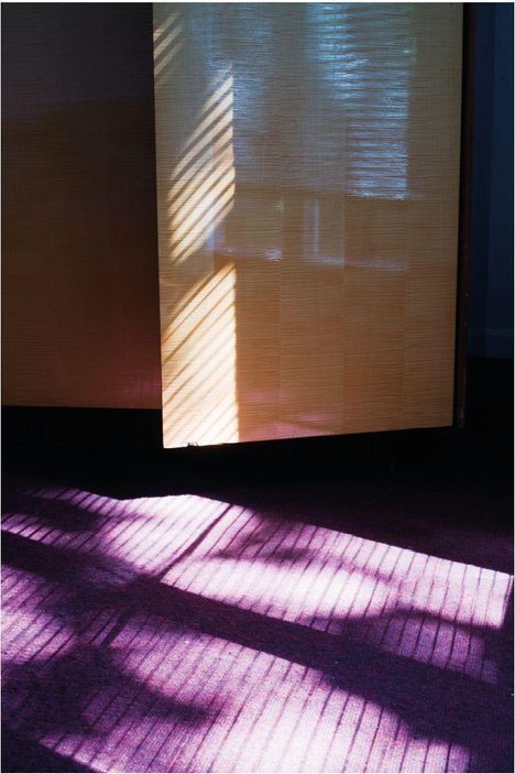Kein Text erkennbar. Sonnenlicht fällt durch Jalousien auf einen gelben Vorhang und wirft Schattenspiel auf lila Teppich.
