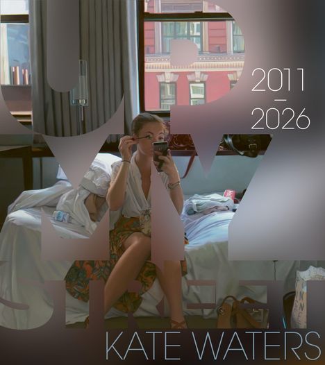 2011-2026, Kate Waters. Eine Frau schminkt sich auf einem Bett, durch Fenster ist ein Gebäude sichtbar.