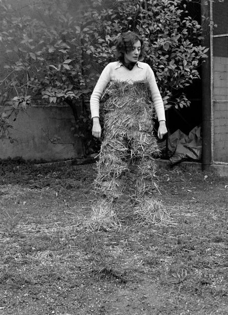 Keine sichtbaren Texte. Eine Person in einem Garten trägt Heu als Kleidungsstück. Im Hintergrund sind Pflanzen zu sehen.