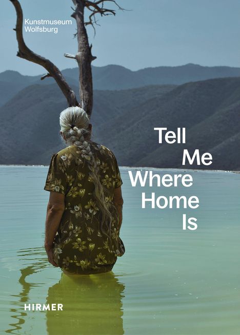 Texte: "Kunstmuseum Wolfsburg," "Tell Me Where Home Is," "HIRMER." Frau steht im Wasser, Baum, Berge im Hintergrund.