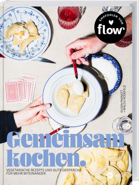 „Gemeinsam kochen. Vegetarische Rezepte und gute Gespräche für mehr Miteinander.“ Zwei Teller mit Teigtaschen, ein Glas Rotgetränk, Spielkarten.
