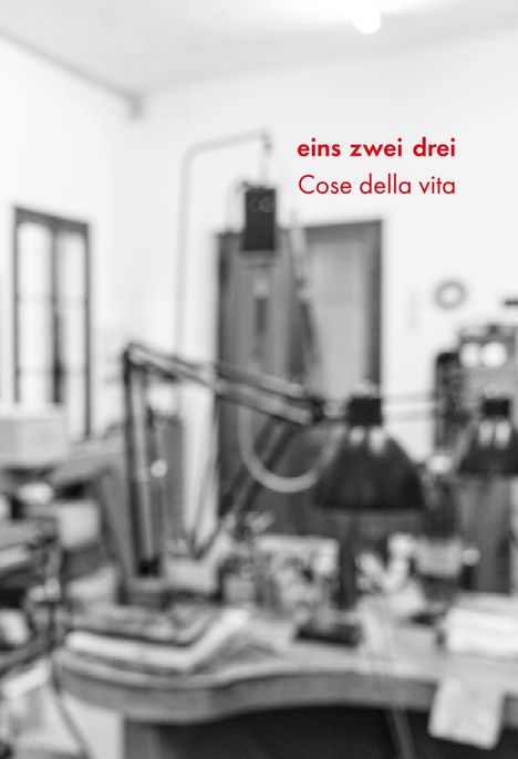 Text: "eins zwei drei, Cose della vita". Unschärfer Raum mit mehreren Schreibtischlampen und Regalen im Hintergrund.