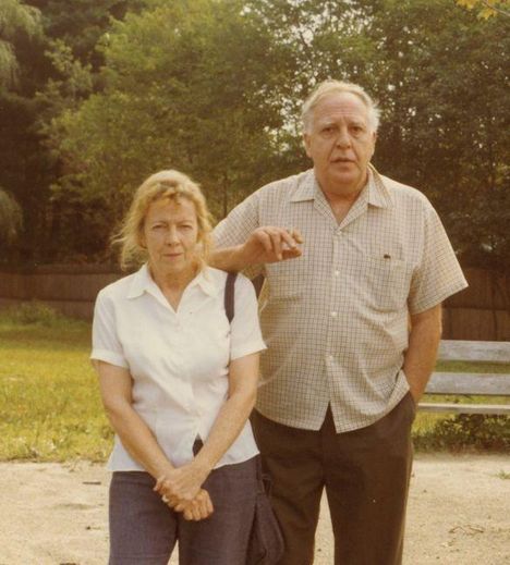 Ein Mann und eine Frau stehen in einem Garten. Der Mann hält eine Zigarre. Bäume sind im Hintergrund sichtbar.