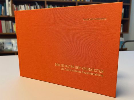 Oranges Buchcover mit dem Titel "DAS ZEITALTER DER KREMATISTEN: 150 Jahre moderne Feuerbestattung". Im Hintergrund Bücherregal.