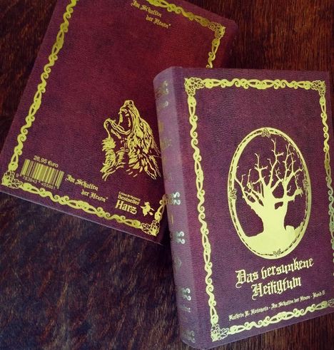 Zwei dunkelrote Bücher mit goldenen Verzierungen. Auf einem steht "Das versunkene Heiligtum", beim anderen ein Wolfskopf.