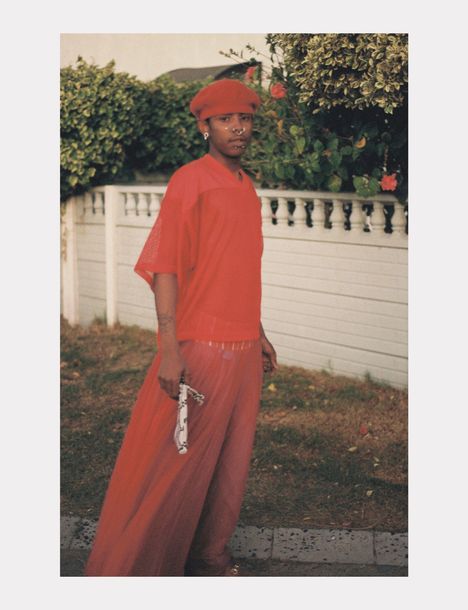 Keine erkennbaren Texte. Eine Person in roter Kleidung und Mütze steht vor einer weißen Mauer, hält ein gemustertes Tuch.