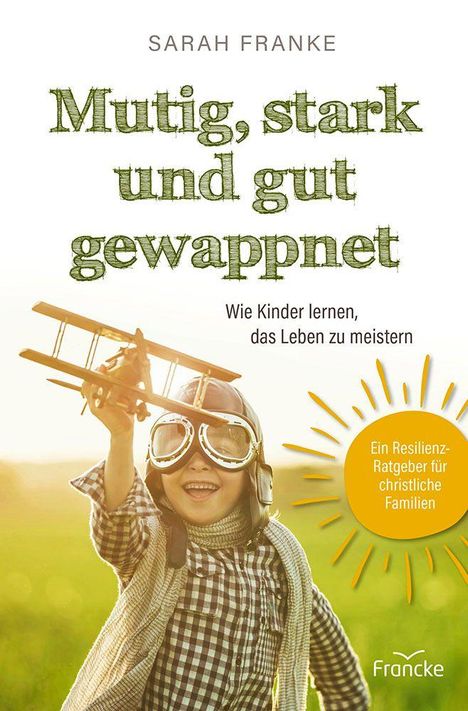 „Mutig, stark und gut gewappnet.“ Ein Junge mit Pilotenbrille hält ein Holzflugzeug in einem Feld hoch.