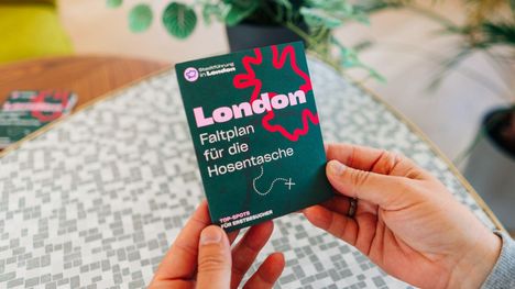 „London Faltplan für die Hosentasche.“ Hände halten eine faltbare Karte. Hintergrund mit Pflanzen und Tischmuster.