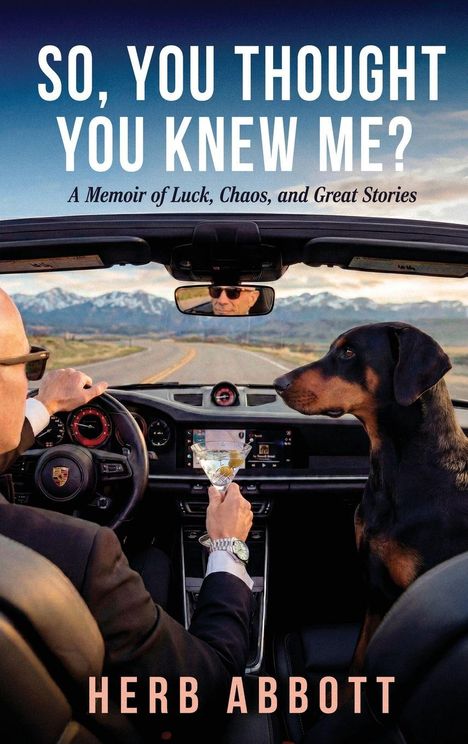 Ein Mann im Cabrio hält einen Cocktail, ein Hund sitzt neben ihm. Beide schauen auf eine Straße mit Bergen im Hintergrund.