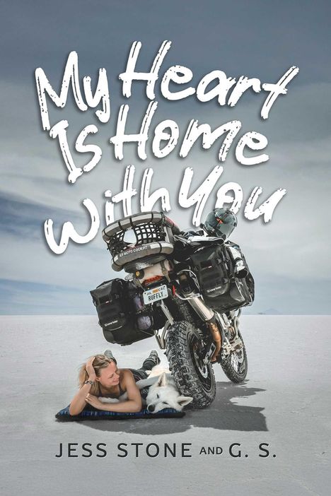 "My Heart Is Home With You" in handschriftlichem Stil. Eine Frau und ein Hund ruhen unter einem Motorrad auf sandigem Boden.