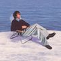 Ein Mann sitzt entspannt in einem Liegestuhl, trägt Sonnenbrille und dunklen Pullover, beobachtet das blaue Meer vor ihm.