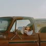 Ein rostiger Pickup mit Text "Custom Deluxe 10". Eine Person lehnt entspannt aus dem Fenster. Steinige Landschaft im Hintergrund.