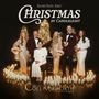„Christmas by Candlelight“. Fünf elegant gekleidete Menschen, umgeben von Kerzen und leuchtenden Bäumen.