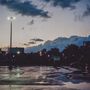 Nasser Parkplatz bei Abenddämmerung, vereinzelte Lichter, wolkiger Himmel und im Hintergrund Silhouetten von Gebäuden.