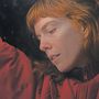 Eine Person mit roten Haaren, die einen roten Mantel trägt. Ihr Gesicht ist nachdenklich und leicht zur Seite geneigt.