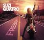 "SUZI QUATRO" und "FREEDOM" sind sichtbar. Eine Frau auf einem Motorrad erhebt die Faust auf einer Wüstenstraße.