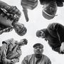 Es sind fünf Männer in einem Kreis, die in die Kamera blicken, teilweise mit Basecaps und Bandanas gekleidet.