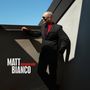 Text: Matt Bianco Masquerader. Ein Mann im Anzug mit Sonnenbrille lehnt an einer Mauer, dramatische Schatten.