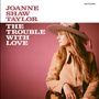 Text: "JOANNE SHAW TAYLOR, THE TROUBLE WITH LOVE." Eine Frau in Hut und Jacke hält eine Gitarre vor rosa Hintergrund.