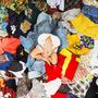 Viele bunte Kleidungsstücke umgeben eine in weiß gekleidete Person, die mit überkreuzten Beinen sitzt.