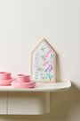 Keine Texte erkennbar. Eine Illustration von Blumen in einem holzgerahmten Hausform mit zwei rosa Kerzenhaltern daneben.