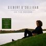 "GILBERT O'SULLIVAN ON THE RECORD. Person sitzt auf einem Hügel am Wasser, im Hintergrund Bäume."