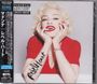 Japanische Schriftzeichen links, "Rebel Heart" auf dem Arm, Frau in rotem Kleid lächelt mit Händen am Gesicht.