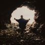Kein Text vorhanden. Silhouette einer Person mit ausgebreiteten Armen steht am Eingang einer felsigen Höhle, Licht im Hintergrund.