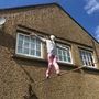 Es gibt keinen sichtbaren Text. Eine Gestalt in pinker Kleidung balanciert auf einem Seil vor einem Haus mit zwei Fenstern.