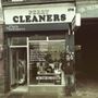 „Perry Cleaners“ steht als großes Schild über einem kleinen Laden. Ein Mann lehnt entspannt an der Fassade.