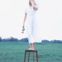 Keine erkennbaren Texte. Eine Person steht barfuß auf einem Hocker in einem weißen Kleid und hält eine Rose in der Hand.
