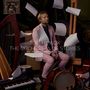 Texte: "TOM HICKOX THE ORCHESTRA OF STORIES FEAT. THE CHINEKE! ORCHESTRA & ONYX BRASS". Ein Mann in rosa Anzug, umgeben von Instrumenten und schwebenden Notenblättern.